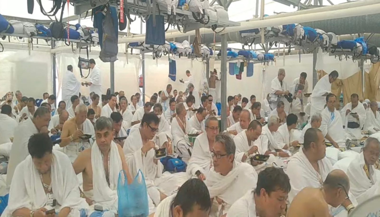 Jemaah haji pria berihram duduk di dalam tenda besar di Mina, beberapa memegang kotak makanan.