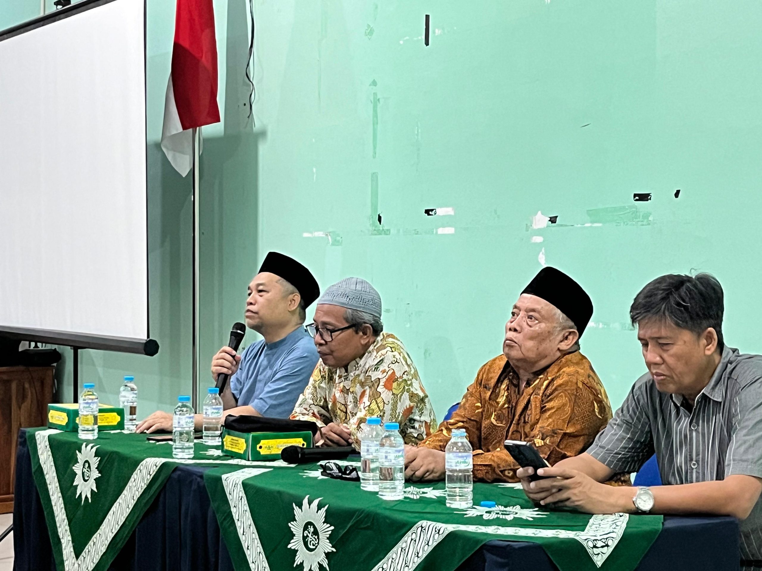Foto dokumentasi rapat koordinasi di sebuah aula besar. Di bagian depan, terdapat meja pimpinan dengan beberapa narasumber pria yang mengenakan batik dan peci hitam. Salah satu pimpinan sedang berbicara di depan mikrofon. Di depan mereka, puluhan peserta pria yang mengenakan seragam batik Muhammadiyah (batik dasar krem dengan motif hijau) duduk rapi di barisan kursi yang memenuhi ruangan. Suasana tampak khidmat dan terorganisir dengan latar belakang spanduk kegiatan di dinding aula.