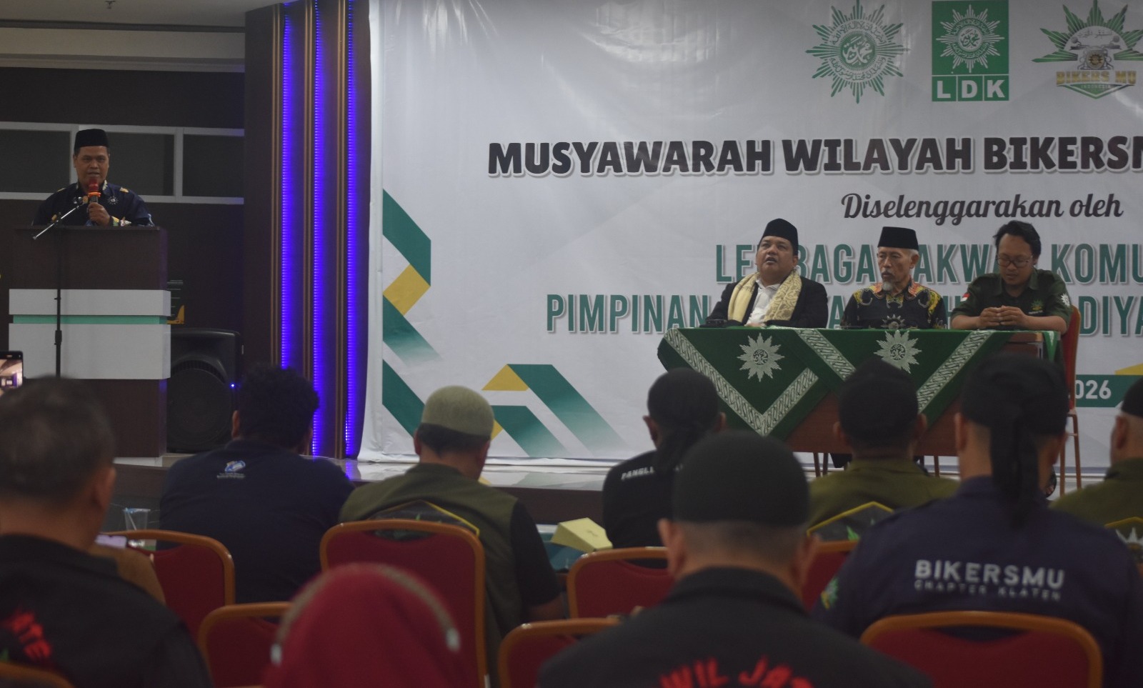 Foto suasana Musyawarah Wilayah (Musywil) BikersMu Jawa Tengah di dalam Aula Rumah Sakit Unimus. Di latar belakang terdapat spanduk besar bertuliskan "MUSYAWARAH WILAYAH BIKERSMU" yang diselenggarakan oleh Lembaga Dakwah Komunitas Pimpinan Wilayah Muhammadiyah. Di atas panggung, terlihat para pimpinan sedang duduk di meja utama, sementara seorang pembicara berdiri di podium sebelah kiri mengenakan seragam hitam. Di bagian depan, tampak deretan kursi merah yang diisi oleh peserta jemaah yang mengenakan jaket seragam hitam bertuliskan "BIKERSMU" dan nama chapter daerah masing-masing.
