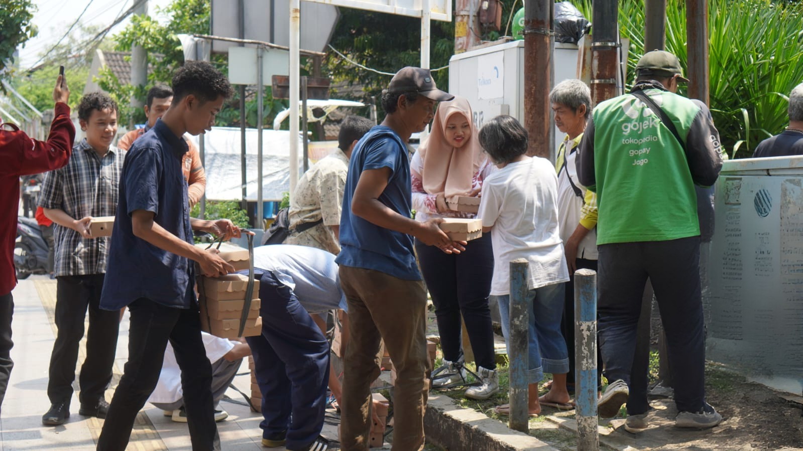 Foto suasana di pinggir jalan raya di Semarang yang menunjukkan sekelompok remaja mengenakan pakaian bebas sedang membagikan paket nasi kotak kepada warga sekitar. Tampak seorang siswa membawa tumpukan kotak makanan berwarna cokelat menggunakan tali pengikat, sementara rekannya yang lain sedang menyerahkan kotak makanan kepada seorang ibu dan pria paruh baya. Di lokasi tersebut juga terlihat seorang pengemudi ojek online yang sedang berdiri membelakangi kamera. Suasana tampak tertib dan dipenuhi interaksi hangat di trotoar jalan yang cerah.