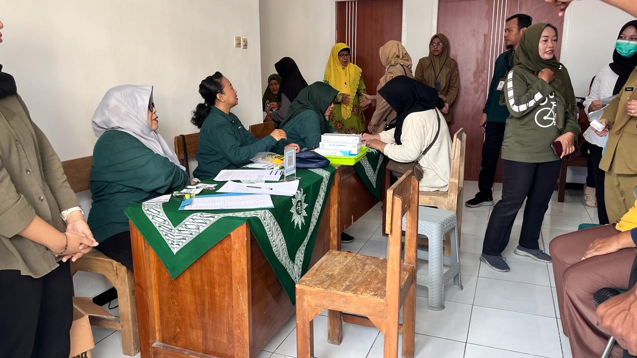 Foto dokumentasi suasana meja pendaftaran di sebuah ruangan pelayanan kesehatan. Di sisi kiri, tampak tiga orang kader wanita 'Aisyiyah mengenakan seragam hijau tua dan jilbab abu-abu sedang melayani proses administrasi warga. Di atas meja kayu yang dilapisi taplak hijau bermotif batik khas Muhammadiyah/'Aisyiyah, terdapat berkas formulir pendaftaran, alat tulis, dan kotak obat-obatan. Di sisi kanan dan latar belakang, tampak beberapa ibu dan petugas puskesmas atau Disdalduk mengenakan seragam dinas cokelat cokelat sedang mengantre dan berkoordinasi, menciptakan suasana pelayanan yang tertib, sibuk, dan ramah.
