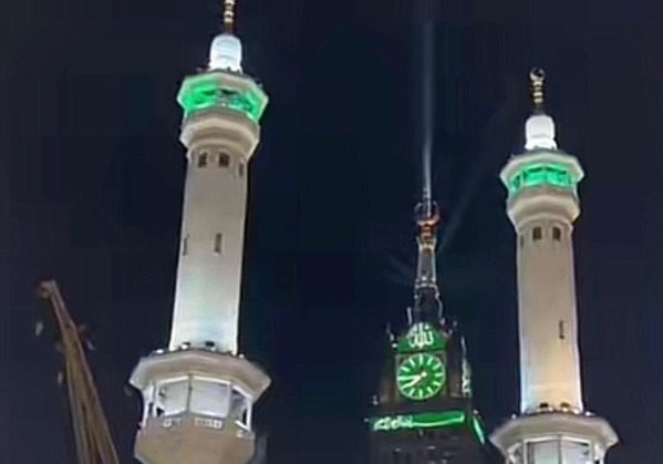 Foto dokumentasi suasana malam hari di Kota Makkah yang menampilkan bagian atas dua menara Masjidil Haram yang megah dengan lampu sorot berwarna hijau di puncaknya. Di antara kedua menara tersebut, tampak puncak Tower Jam Zam-Zam yang ikonik memancarkan kilauan cahaya hijau yang indah ke langit malam. Bagian jam dinding besar pada menara juga menyala terang dengan warna hijau, menciptakan atmosfer spiritual yang syahdu dan menandakan kondisi makkah terkini yang telah memasuki bulan Zulhijah.