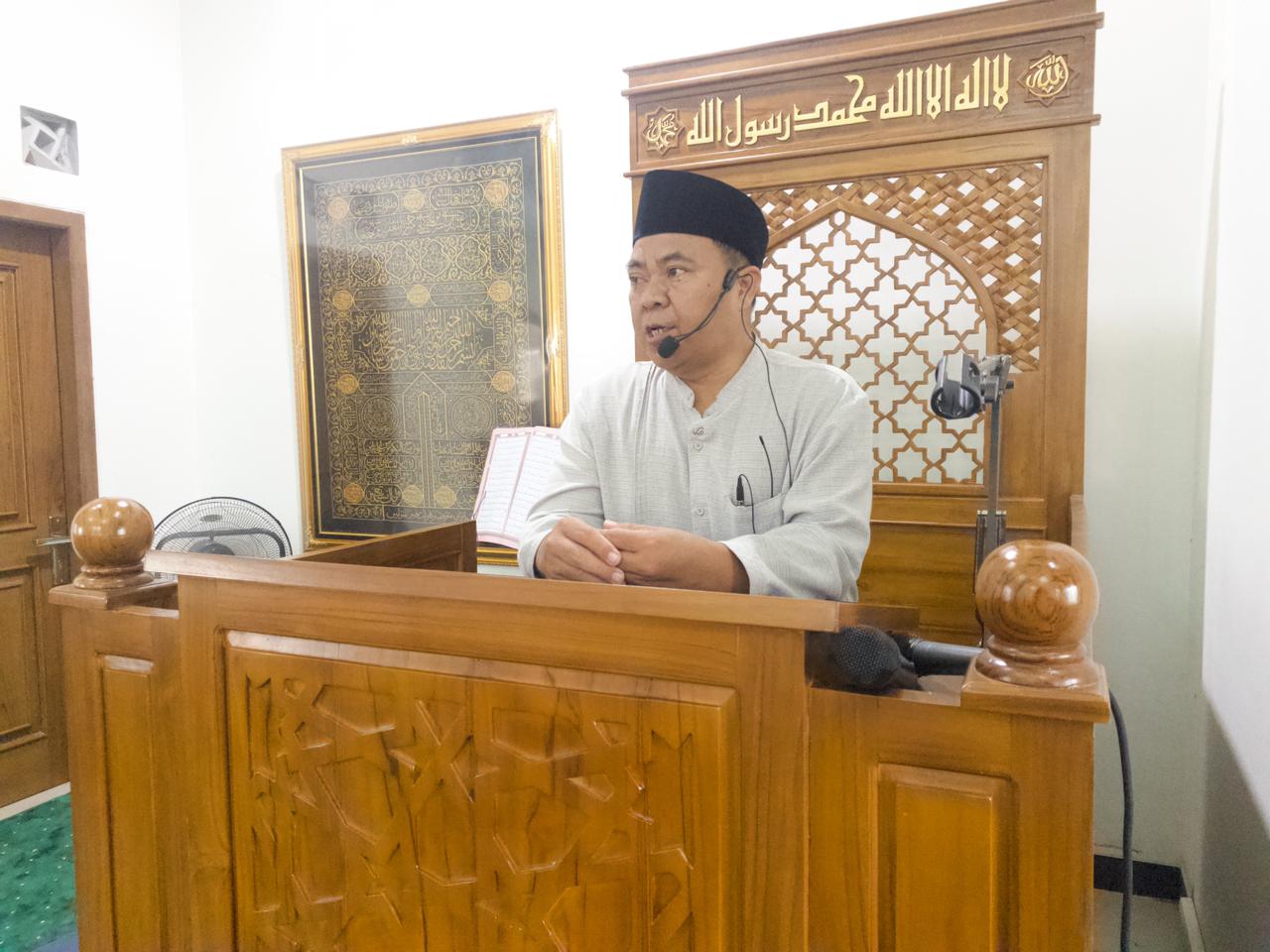 Foto Wakil Ketua PWM Jawa Tengah Dr. H. Ahmad Hasan Asy’ari Ulama’i, M.Ag. sedang menyampaikan khutbah Jumat dari atas mimbar kayu ukir di Masjid At-Taqwa Ngaliyan, Semarang. Beliau mengenakan baju koko putih, peci hitam, dan mikrofon bando (headset mic). Di latar belakang dinding putih, tampak hiasan kaligrafi Arab berbingkai emas yang estetik.