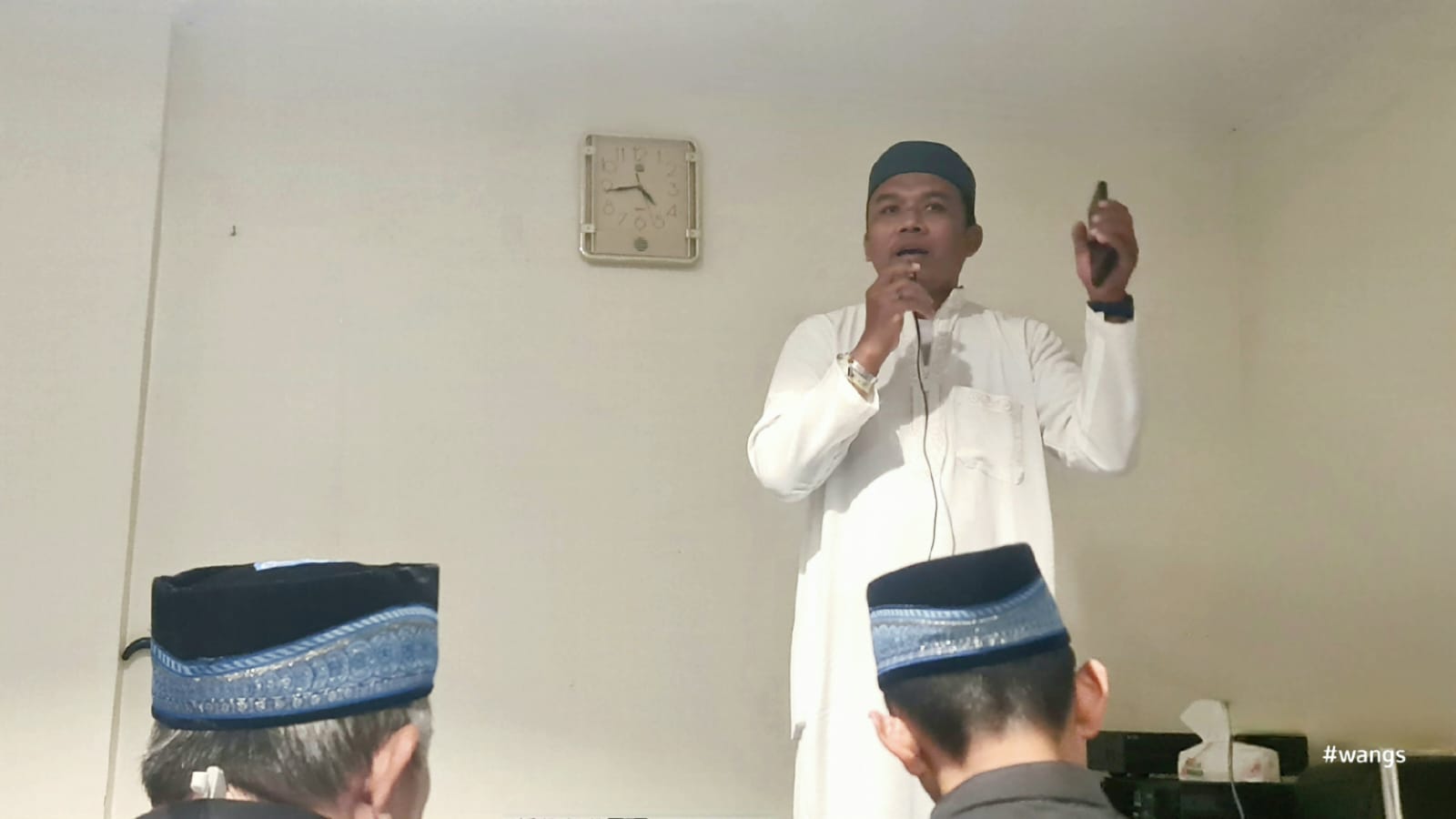 Foto Ustaz Amin Thoha berdiri memberikan kultum subuh dengan pakaian gamis putih dan peci hitam di dalam ruangan hotel jemaah haji di Makkah. Tangan kanannya memegang mikrofon dekat bibir, sementara tangan kirinya sedikit terangkat memegang gawai. Di bagian latar depan bawah, tampak bagian belakang kepala dua orang jemaah pria yang mengenakan peci hitam bermotif biru sedang menyimak ceramah.