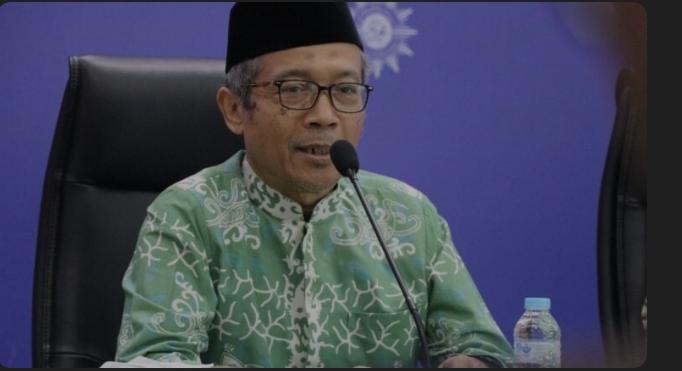Foto dokumentasi setengah badan almarhum Prof. Hamim Ilyas sedang berbicara dengan tenang di depan mikrofon dalam sebuah forum resmi Muhammadiyah. Beliau mengenakan kacamata, peci hitam, dan kemeja batik berwarna hijau bermotif putih dengan latar belakang biru tua berlogo matahari Muhammadiyah. Foto kenangan ini dirilis bersamaan dengan kabar duka mengenai wafatnya beliau pada Sabtu, 23 Mei 2026.