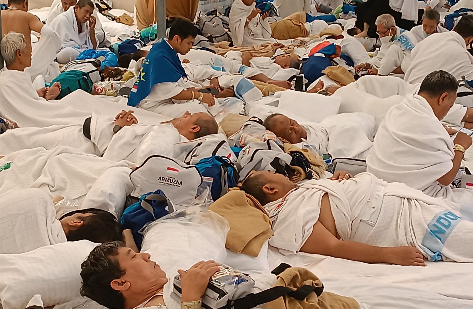 Foto dokumentasi suasana di dalam tenda Mina yang padat, di mana para jemaah haji laki-laki Indonesia yang mengenakan kain ihram sedang beristirahat dan tidur berjejer rapat di atas kasur tipis. Tampak beberapa jemaah meletakkan perlengkapan tas bertuliskan "Armuzna" dan syal bertuliskan "INDONESIA", menggambarkan ketahanan fisik jemaah setelah menempuh perjalanan jauh untuk menunaikan ibadah tarwiyah