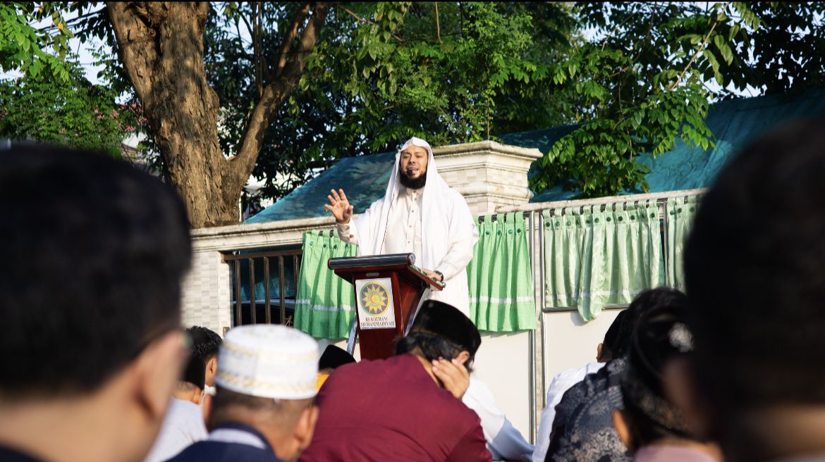 Foto dokumentasi Syekh Hamdi Al Misri yang mengenakan jubah putih bersih dan penutup kepala tradisional Timur Tengah senada sedang berkhutbah dengan mantap dari atas mimbar kayu. Tangan kanan beliau tampak terangkat dengan telapak terbuka untuk menekankan pesan khutbahnya. Di barisan depan bawah, tampak bagian belakang jemaah pria berkopiah dan berpeci yang sedang duduk bersila menyimak materi secara khusyuk saat pelaksanaan shalat Idul Adha 2026 di halaman luar ruangan yang teduh.