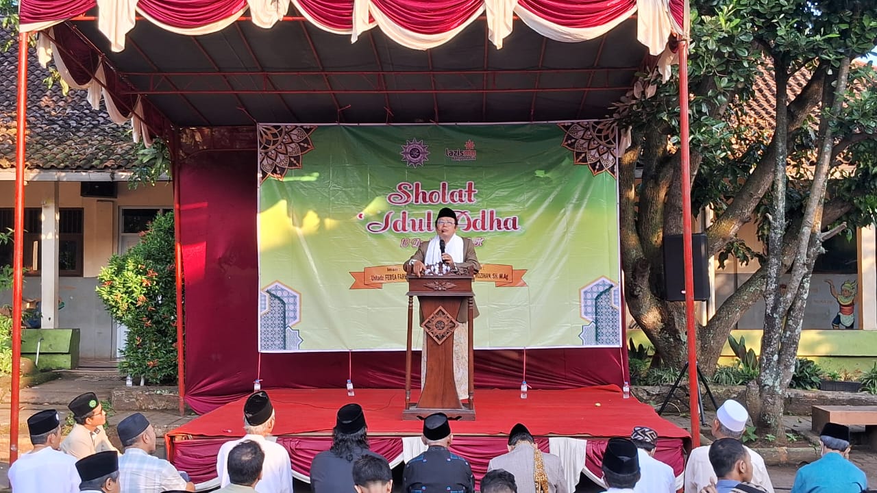 Prof. Dr. Rosihan, M.Ag. sedang berdiri menyampaikan khotbah Iduladha dengan pengeras suara di atas panggung mimbar luar ruangan, berlatar belakang spanduk hijau bertuliskan Sholat Idul Adha PCM Banyumanik, di hadapan jemaah yang duduk menyimak.