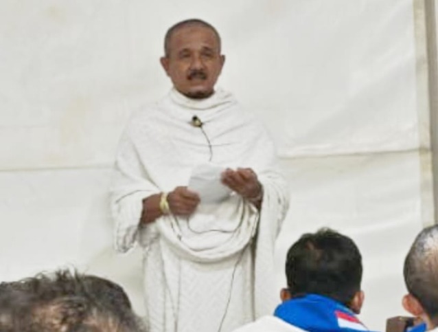 Drs. H. Nurbini, M.Si. mengenakan pakaian ihram putih sedang berdiri menyampaikan khotbah wukuf sambil memegang kertas catatan di hadapan jemaah haji Kloter 28 SOC Semarang di dalam tenda Arafah.