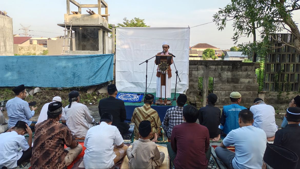 Ustaz Eko Mustofa sedang berdiri tegak menyampaikan khotbah Iduladha dari atas mimbar kayu berornamen matahari Muhammadiyah di halaman terbuka Kelurahan Pandean Lamper, Gayamsari, Semarang, dihadiri oleh barisan jemaah pria yang duduk mendengarkan dengan khusyuk.