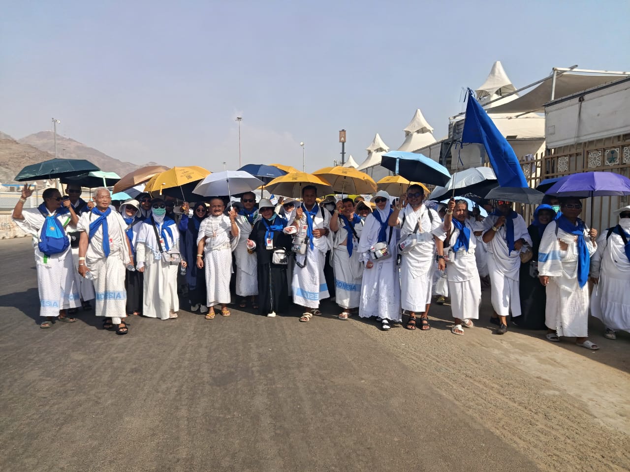 Rombongan besar jemaah haji Indonesia mengenakan pakaian ihram dan mukena putih dengan syal biru di leher, berjalan kompak beriringan di jalan aspal yang luas sambil memegang payung warna-warni di bawah langit cerah.