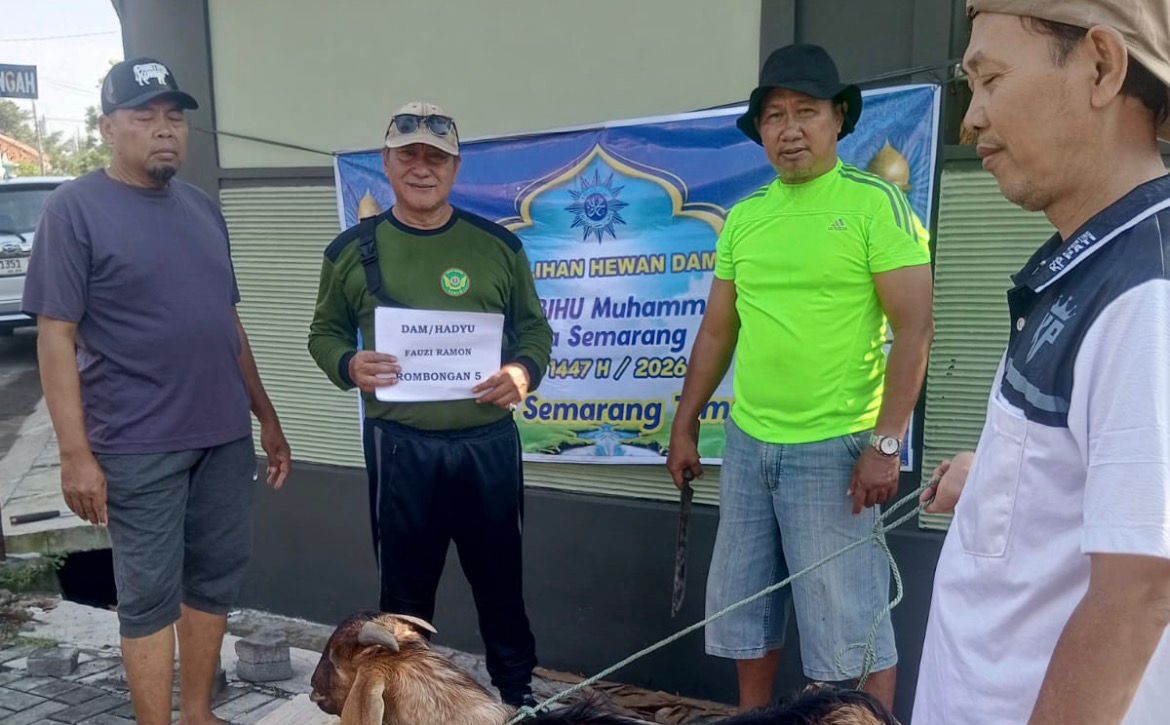 Foto penyerahan hewan Dam/Hadyu di depan spanduk KBIHU Muhammadiyah Kota Semarang. Anggota KOKAM Semarang Timur berbaju navy dan Edi KOKAM pembawa kertas nama jemaah berdiri di sisi kiri. Di tengah, Imam Abu Maulana berkaos hijau terang memegang tali, sementara seorang kader NU berkaos putih membawa kambing kurban di sisi kanan.