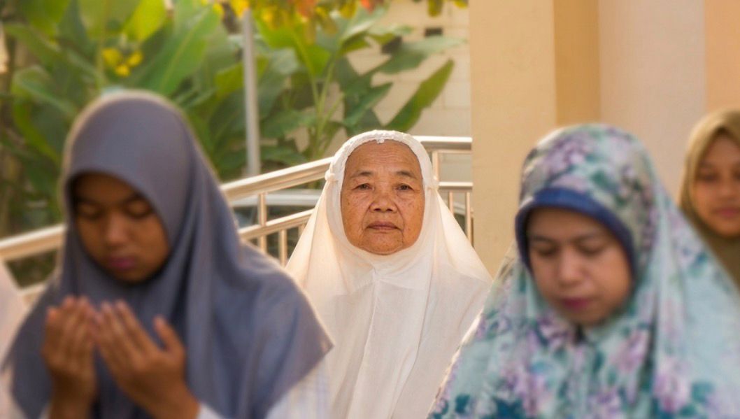 Seorang lansia, Ibu Badriyah (tengah), mengenakan kerudung putih, tampak haru dan damai di antara jemaah wanita lainnya (yang terlihat lebih muda) di area luar Masjid At-Taqwa, Ngaliyan, Semarang. Lansia itu menghadap kamera sementara jemaah di dekatnya menunduk dan berdoa. Cahaya alami lembut menerangi scene.