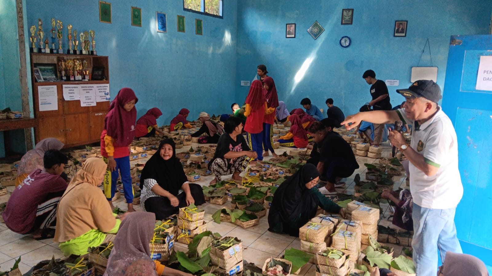 Suasana di dalam ruangan yang dipenuhi panitia kurban laki-laki, perempuan, dan anak-anak sedang sibuk bergotong royong mengemas potongan daging kurban ke dalam wadah besek bambu tradisional beralaskan daun hijau di lantai kelas.