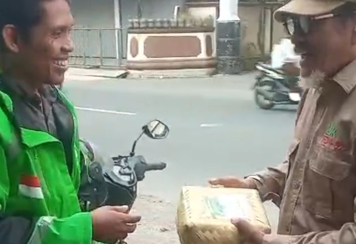 Seorang relawan LDK PDM Kota Semarang mengenakan kemeja cokelat, topi, dan kacamata hitam sedang menyerahkan wadah besek bambu berisi daging kurban kepada seorang pengemudi ojek online berjaket hijau yang tersenyum lebar di pinggir jalan.