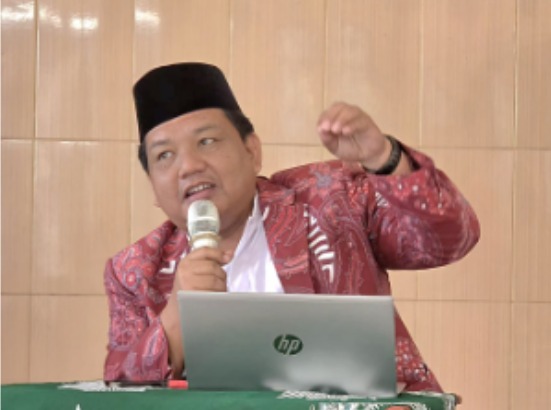 Foto dokumentasi yang menampilkan Dr. AM. Jumai sedang berdiri di belakang podium kayu di dalam sebuah masjid. Beliau mengenakan kemeja batik bermotif warna cokelat dan krem, peci hitam, serta kacamata. Tangan kanan beliau tampak memegang mikrofon saat memberikan penjelasan, sementara tangan kirinya berada di atas meja podium. Di latar belakang, terlihat jemaah pria yang duduk menyimak kajian dengan khidmat di dalam ruangan masjid yang luas.
