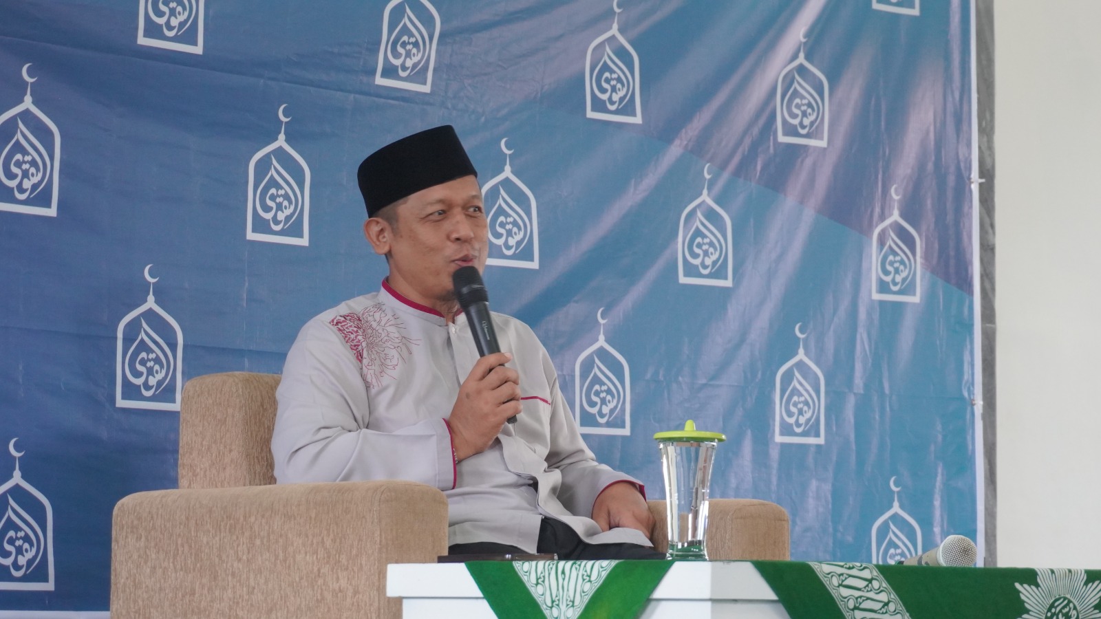 Foto dokumentasi suasana pengajian Ahad pagi di dalam sebuah masjid yang luas dan bersih. Di bagian depan, seorang pimpinan pengajian atau narasumber mengenakan kemeja batik bermotif cokelat, peci hitam, dan kacamata, sedang berbicara menggunakan mikrofon di podium. Di hadapannya, ratusan jemaah pria dan wanita duduk dengan khidmat di atas karpet masjid berwarna hijau, menyimak penyampaian materi dengan tertib, menciptakan atmosfer kajian yang tenang dan religius.