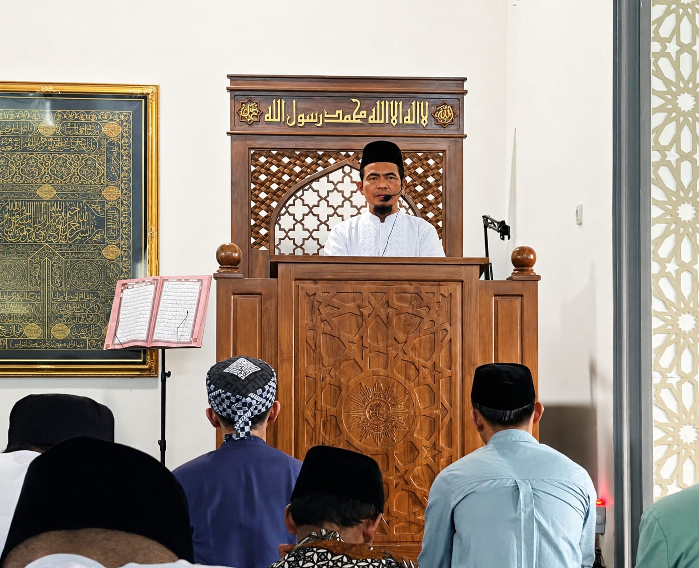 Foto suasana khotbah Jumat di dalam masjid yang menunjukkan Ustadz Ananto Tri Prasetia, S.Pd., sedang berdiri di atas mimbar kayu berwarna cokelat tua yang memiliki ukiran matahari khas Muhammadiyah di bagian depannya. Beliau mengenakan baju koko putih dan peci hitam, berbicara di depan mikrofon. Di sebelah kiri mimbar, tergantung sebuah bingkai besar berisi kaligrafi berwarna emas dengan latar belakang hitam, serta sebuah Al-Qur'an yang terbuka di atas rehal besi. Di bagian depan mimbar, tampak barisan jemaah yang duduk khidmat, termasuk seorang pria mengenakan penutup kepala bermotif batik kotak-kotak.