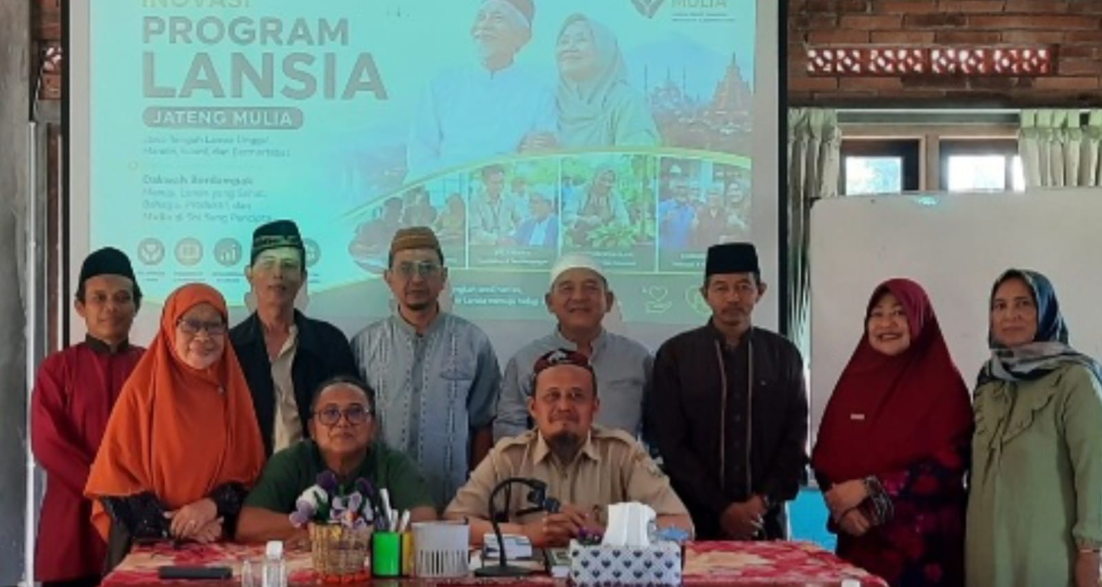 Foto dokumentasi yang menampilkan Fajar Arifin sedang berdiri di sebuah area terbuka dengan latar belakang perbukitan hijau yang asri. Beliau mengenakan kemeja batik bermotif cokelat dan krem, kacamata, serta peci hitam. Wajahnya menunjukkan ekspresi ramah dengan senyum tipis. Di belakangnya tampak pemandangan lereng gunung dengan pepohonan rimbun dan kabut tipis, menciptakan suasana yang tenang, sejuk, dan inspiratif.