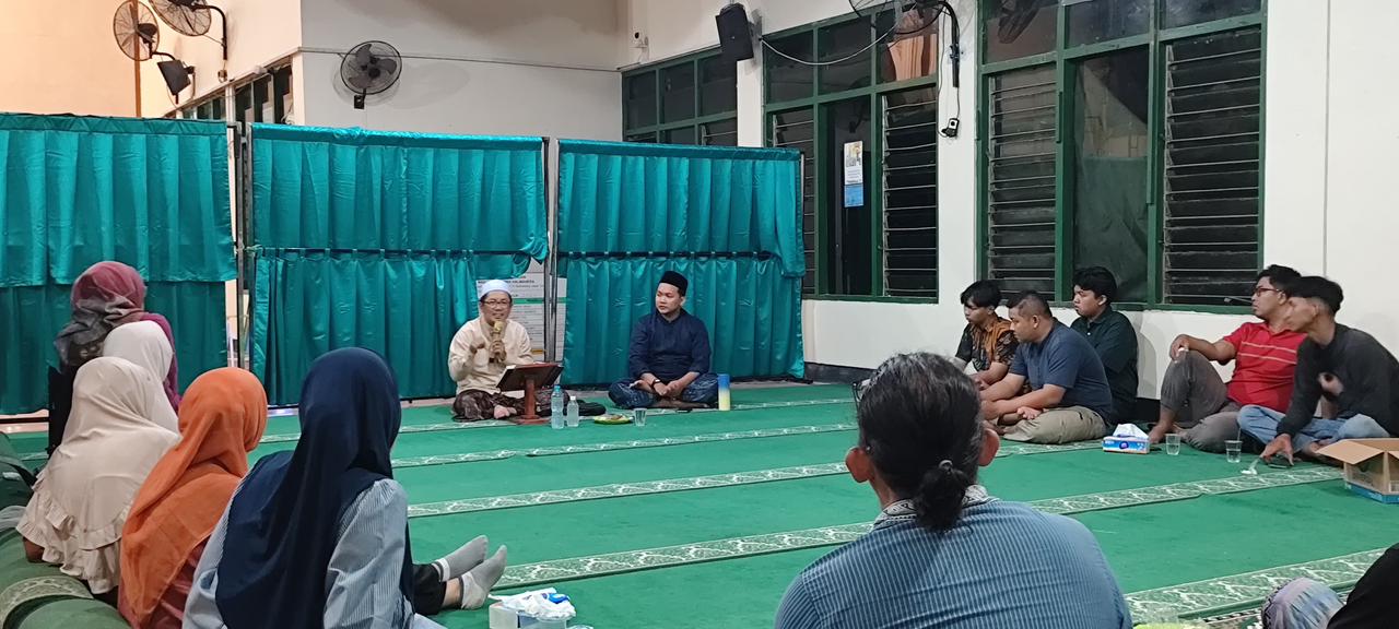 Foto suasana kajian persiapan Iduladha di dalam Masjid Taqwa Halmahera. Terlihat sekelompok jemaah laki-laki dan perempuan duduk bersila di atas karpet hijau sambil menyimak penjelasan materi. Di depan jemaah, seorang ustadz berbaju koko krem dan peci putih sedang berbicara menggunakan mikrofon dengan sebuah rehal buku di hadapannya. Di belakang ustadz terdapat tirai pembatas hijau tua dan jendela kaca besar yang memperlihatkan suasana ruangan masjid yang terang.