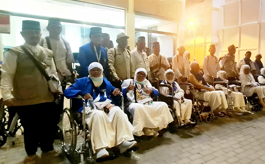 Sekelompok jemaah haji wanita lansia dari Semarang duduk di kursi roda dalam barisan, mengenakan pakaian ihram putih dan tersenyum, dengan petugas berdiri di belakang mereka pada malam hari.