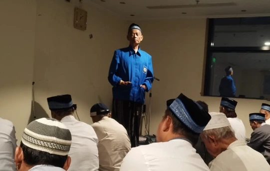 Seorang pria mengenakan rompi biru bertuliskan 'MUHAMMADIYAH' sedang memberikan kultum menggunakan mikrofon di hadapan jemaah haji pria yang duduk lesehan mendengarkan dengan khidmat di sebuah ruangan hotel.