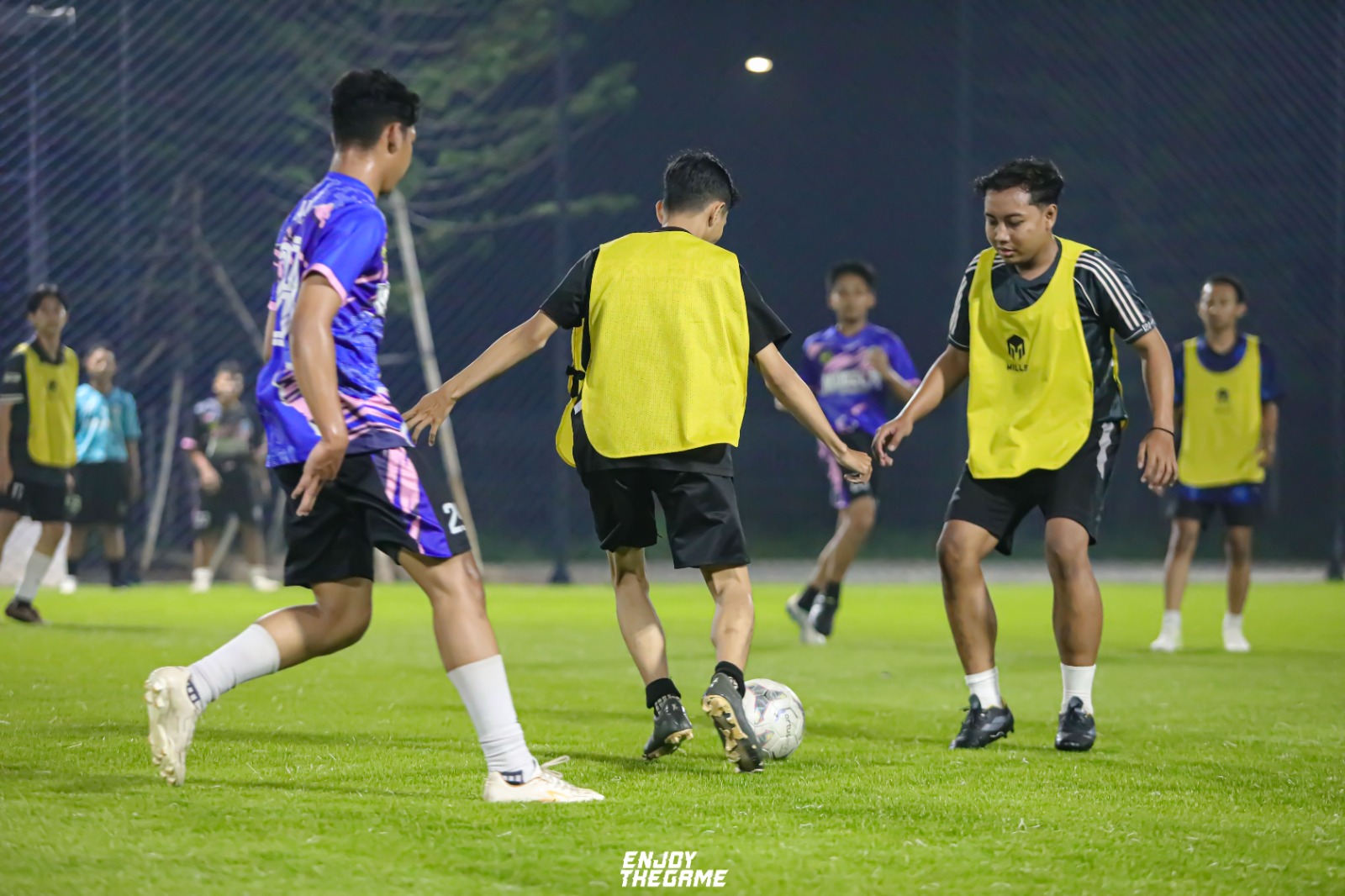 Foto dokumentasi suasana pertandingan mini soccer kasual yang diadakan di Lapangan Up Town Mini Soccer BSB Semarang pada sore hari yang cerah. Tampak beberapa pemuda mengenakan rompi olahraga merah muda (pink) di atas kaus mereka sedang aktif mengejar dan mengoper bola di atas lapangan rumput sintetis hijau yang bersih. Di latar belakang, terlihat jaring pembatas lapangan yang tinggi, pepohonan hijau yang asri, serta beberapa peserta lain yang berdiri di pinggir lapangan menyaksikan jalannya pertandingan dengan antusias, menciptakan atmosfer yang penuh energi, sportivitas, dan kebersamaan.