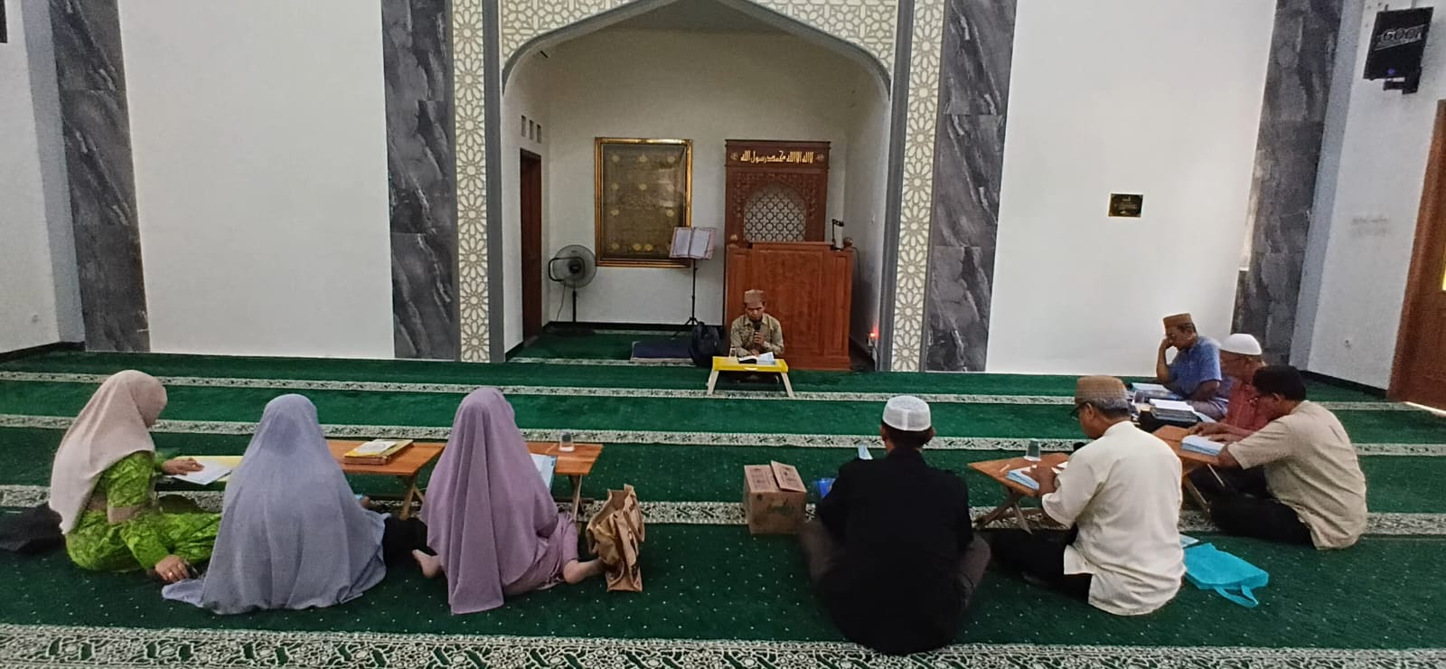 Foto suasana majelis belajar tahsin al quran sore hari di dalam Masjid At-Taqwa Wates, Ngaliyan, Semarang. Tampak Ust. Ananto Tri Prasetia selaku pembimbing duduk di depan mihrab menghadap meja kecil. Di depannya, para jemaah pria dan wanita duduk lesehan terpisah di atas karpet hijau dengan meja-meja kayu kecil untuk meletakkan mushaf Al-Qur'an baru mereka.
