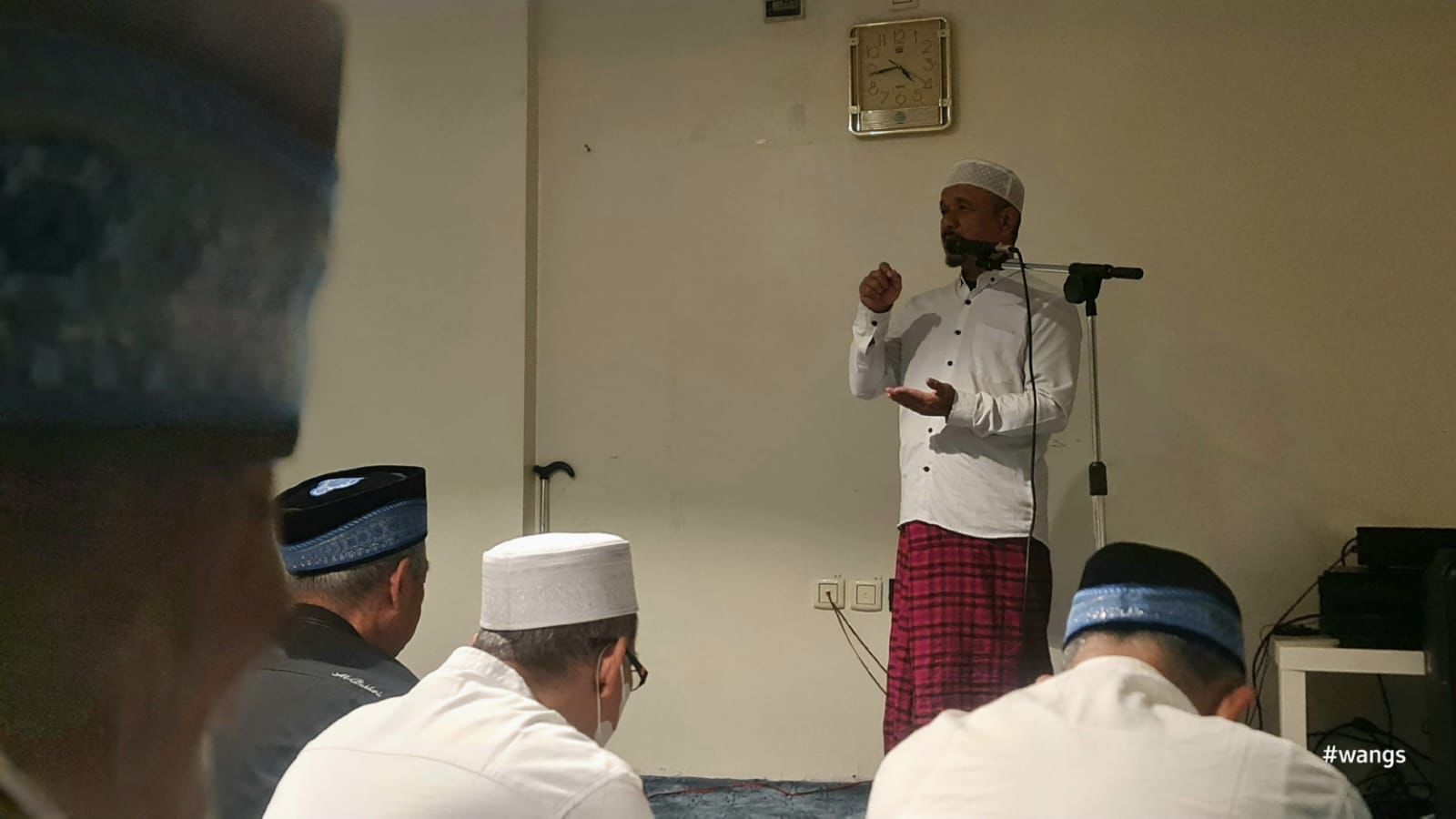 Foto dokumentasi suasana kultum bakda subuh di dalam ruangan akomodasi jemaah haji Indonesia. Di bagian depan, Drs. H. Nurbini, M.S.I. berdiri tegak mengenakan kemeja koko putih lengan panjang, sarung bermotif kotak-kotak merah, dan peci rajut putih. Tangan kanannya tampak mengepal ke atas dan tangan kirinya terbuka saat berbicara di depan mikrofon stand, memberikan penekanan pada materi agar jemaah siap menghadapi ujian fisik karena ruang tenda mina terbatas. Di hadapannya, beberapa jemaah laki-laki mengenakan pakaian putih dan peci hitam serta biru sedang duduk bersila di atas karpet, mendengarkan penjelasan dengan khusyuk.