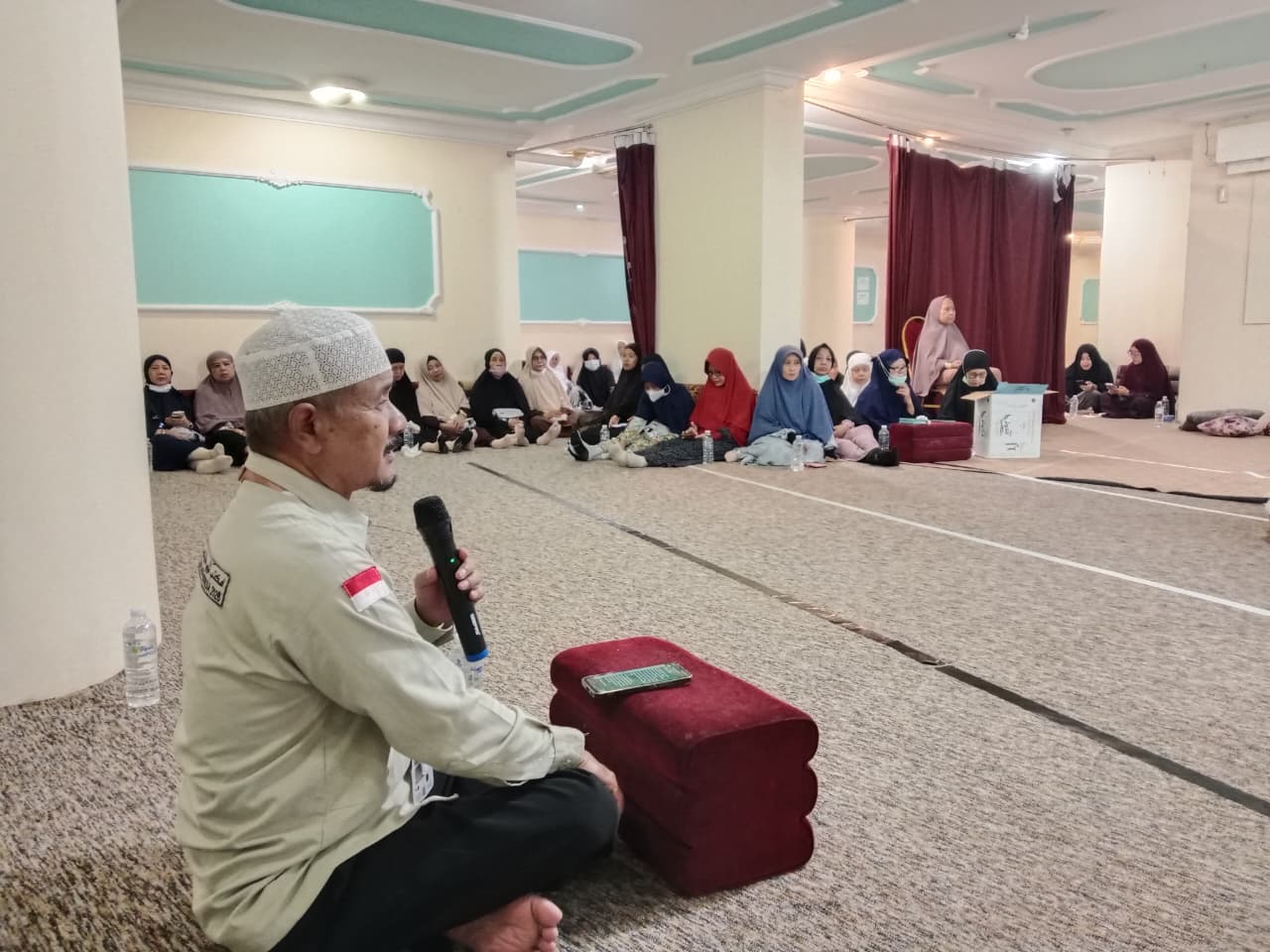 Foto dokumentasi Ustadz Drs. H. Nurbini, M.S.I. sedang memberikan kajian bimbingan ibadah haji mengenai makna hari tarwiyah di aula Darb Almotamer Hostel 2, Syishah, Makkah. Tampak Ustadz Nurbini duduk bersila menghadap kamera dari samping kiri sambil memegang mikrofon hitam. Di latar belakang, puluhan jemaah haji perempuan KBIHU Aisyiyah Kota Tegal duduk lesehan dengan khusyuk mendengarkan materi di dalam aula hotel yang luas dan berkarpet.