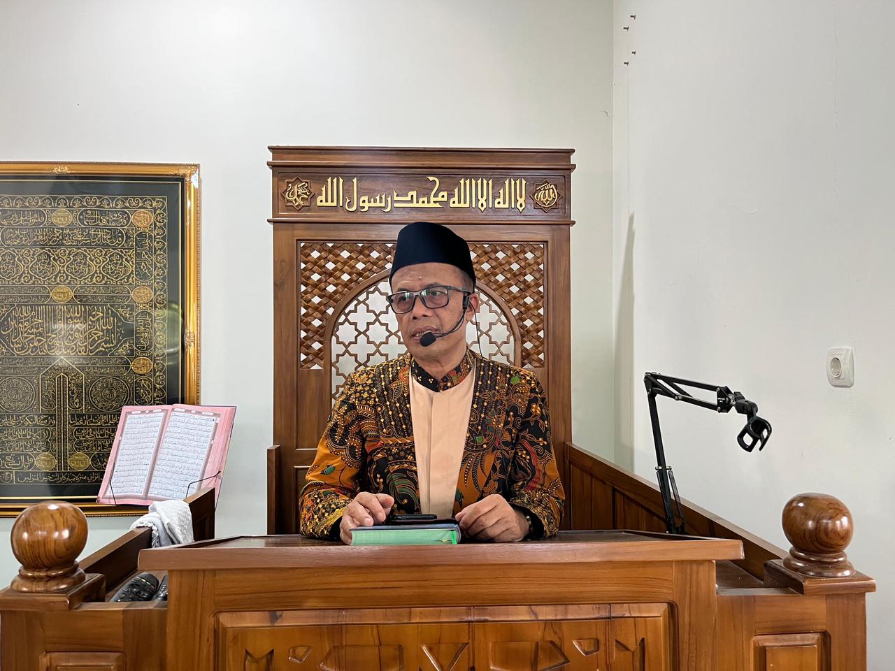 Foto yang menampilkan H. Suparno, S.Ag., M.Si. sedang duduk di balik mimbar kayu berwarna cokelat tua yang memiliki ukiran kaligrafi Arab di bagian atasnya. Beliau mengenakan kemeja batik bermotif dominan oranye dan cokelat, peci hitam, serta kacamata. Di depan beliau terdapat sebuah kitab hijau. Di latar belakang sebelah kiri, tampak sebuah bingkai besar berisi kaligrafi berwarna emas dengan dasar hitam, menciptakan suasana ruang ibadah yang khidmat dan religius