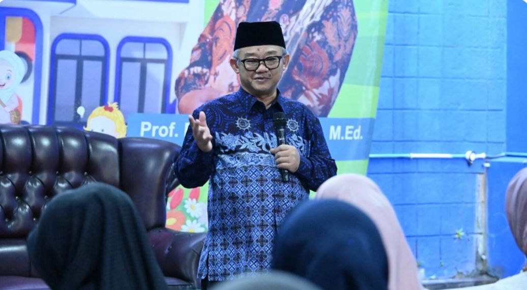 Foto Abdul Mu’ti sedang berdiri mengenakan baju batik lengan panjang bermotif dominan biru tua dan peci hitam. Beliau tampak sedang memberikan paparan dengan memegang mikrofon di tangan kiri dan tangan kanan yang terbuka menunjukkan gestur penjelasan. Di latar belakang, terdapat spanduk atau baliho besar yang menampilkan foto beliau dan tulisan nama serta gelar akademis. Di bagian depan, terlihat bagian belakang kepala beberapa jemaah atau peserta yang sedang menyimak materi secara khidmat.