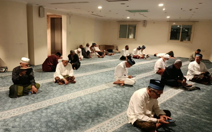 Jemaah Haji Indonesia kloter 28 SOC duduk bersila di karpet hijau musala penginapan membaca Al-Qur'an dan berzikir.