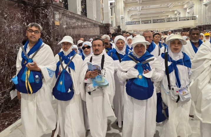 Sekelompok jemaah haji Indonesia dari depan, mengenakan ihram, dipimpin seorang pria memegang buku doa di dalam koridor Masjidil Haram, Makkah.