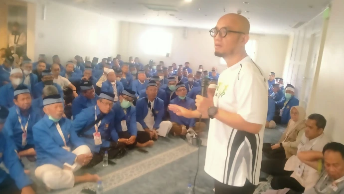 Wamenhaj Dahnil Anzar Simanjuntak memberikan sambutan di depan jamaah haji Kloter 28 SOC Semarang di hotel pemondokan. Beliau memegang mikrofon dan berbicara, sementara jamaah duduk di lantai mengenakan seragam biru dan peci.