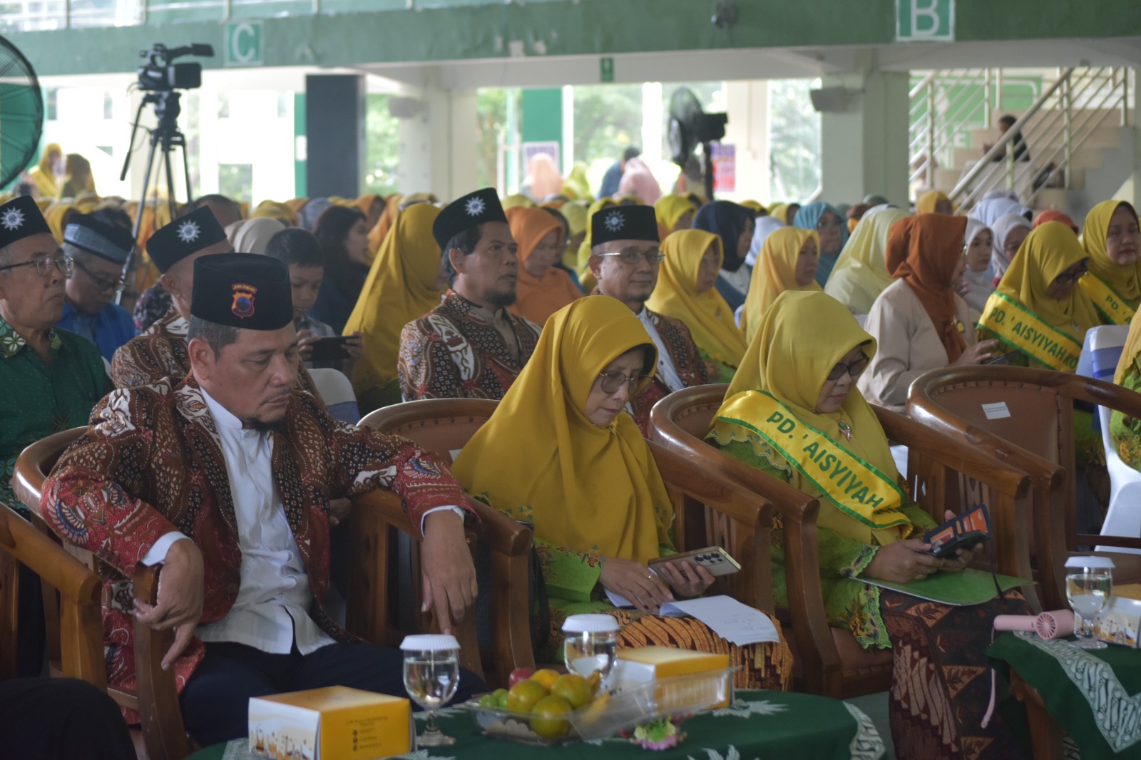 Foto dokumentasi suasana khidmat barisan pimpinan dan tamu undangan yang duduk di barisan depan ruang pertemuan Gedung Serba Guna UNIMUS. Tampak seorang tokoh pria di sisi kiri mengenakan jas batik bercorak cokelat-merah dan peci hitam dengan emblem resmi Jawa Tengah di bagian depan. Di sebelahnya, duduk dua tokoh perempuan penting Aisyiyah berkerudung kuning khas persyarikatan; tokoh di tengah mengenakan kacamata dan gamis hijau, sedangkan tokoh di kanan mengenakan selempang kuning bertuliskan "PD. 'AISYIYAH K..." berwarna hijau. Di latar belakang, ratusan jemaah perempuan berkerudung kuning duduk dengan tertib menyimak jalannya acara di bawah balkon hijau gedung olahraga.