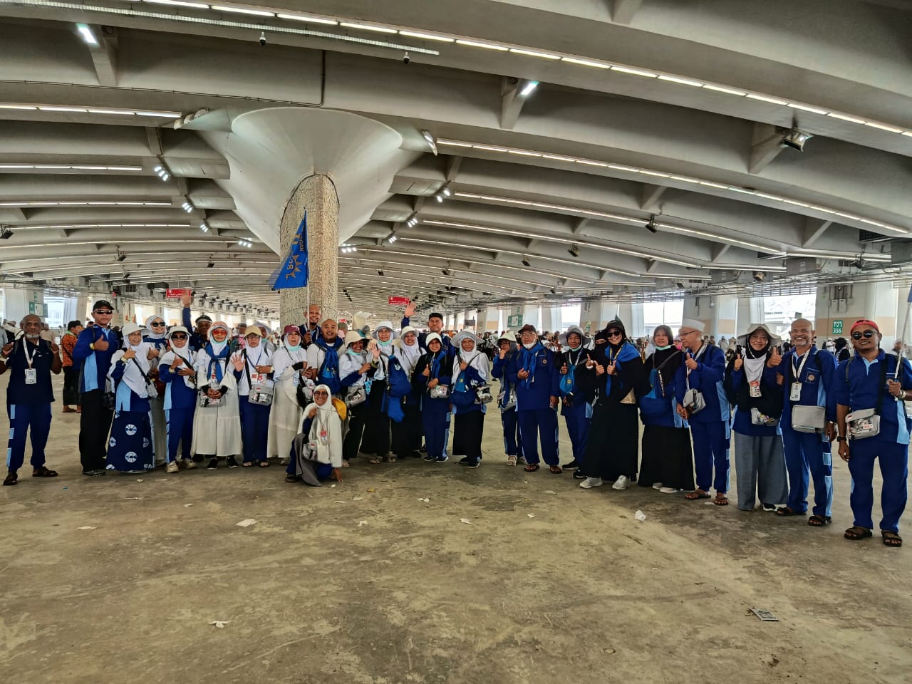 Rombongan besar jemaah haji asal Semarang dengan seragam biru kombinasi putih berfoto bersama dengan ceria di bawah naungan area beratap beton jalur Jamarat, beberapa jemaah tampak melambaikan tangan dan mengacungkan jempol ke arah kamera.