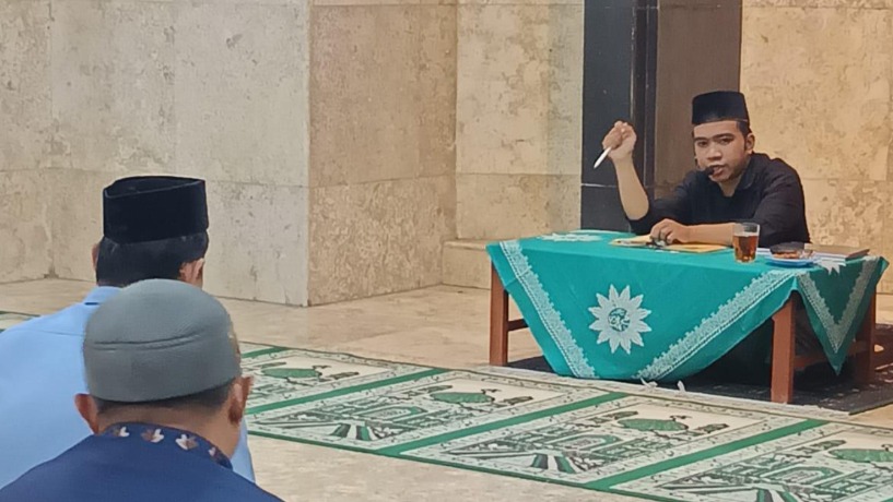 Ustadz M Burhan Khaerudin sedang menyampaikan materi kajian fikih waqaf dengan memegang pena di atas meja belajar berbalut taplak hijau berlogo matahari Muhammadiyah, disimak oleh jemaah pria dari arah belakang.