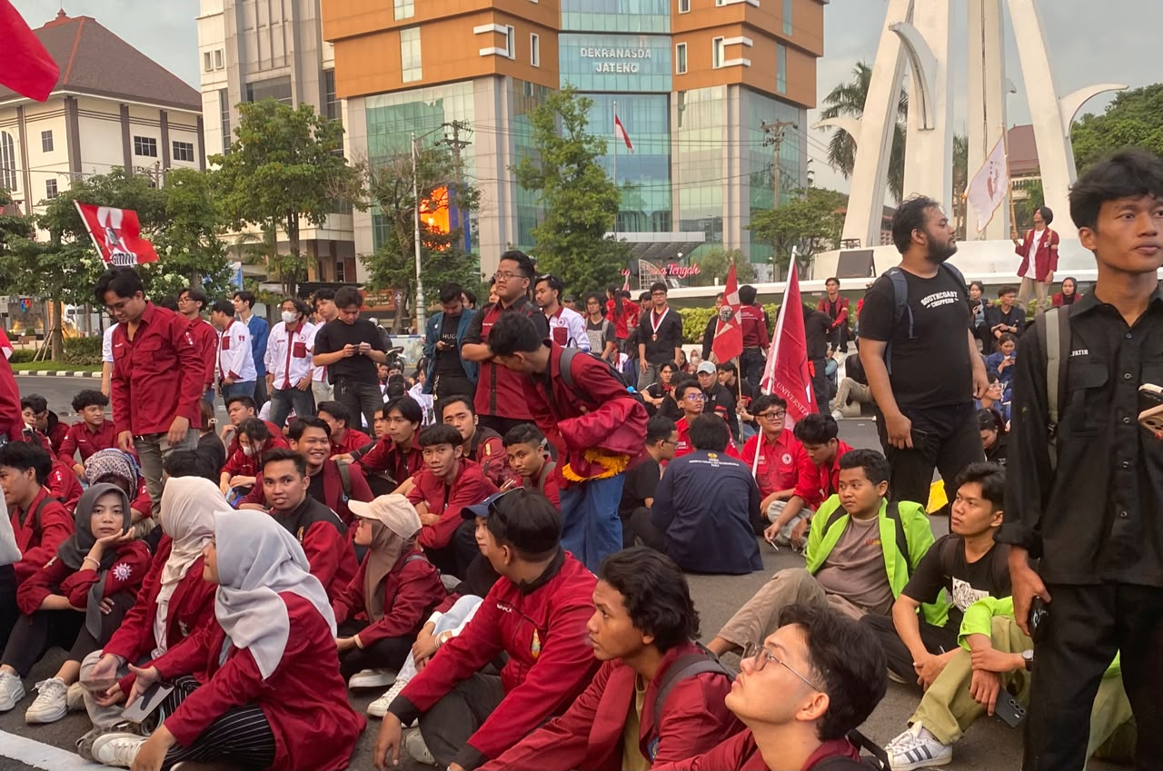 Foto dokumentasi suasana demonstrasi di mana puluhan mahasiswa berpakaian jas almamater merah marun khas IMM sedang duduk tertib merapat di atas aspal Jalan Pahlawan depan Kantor Gubernur Jawa Tengah. Tampak beberapa bendera merah berlambang IMM berkibar di area aksi, dengan latar belakang gedung tinggi Dekranasda Jateng yang menjulang di bawah langit sore yang berawan.