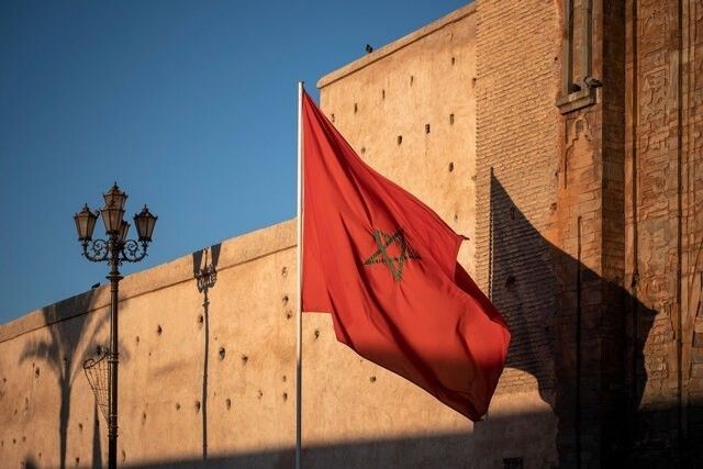 Foto keindahan bendera merah Maroko berlogokan bintang hijau segi lima berkibar gagah di tiang putih, berlatar belakang bangunan dinding benteng kuno berwarna cokelat keemasan di bawah langit biru yang bersih di bawah paparan sinar matahari sore.