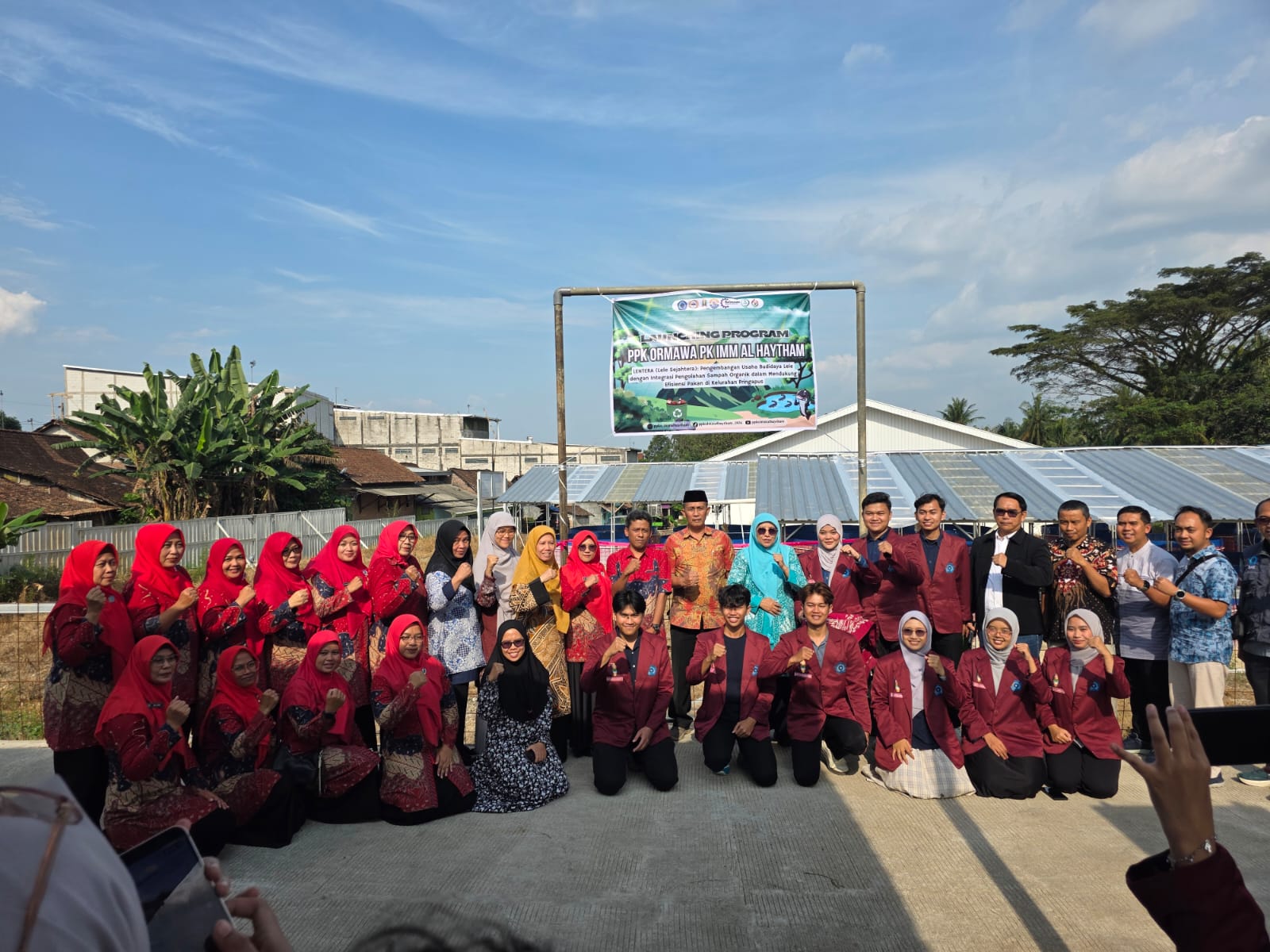 Foto bersama puluhan peserta peluncuran program LENTERA di Kelurahan Pringapus. Di sebelah kiri tampak barisan ibu-ibu PKK mengenakan seragam batik dan jilbab merah menyala. Di bagian tengah berdiri jajaran pimpinan Unimus mengenakan batik dan mahasiswa PK IMM Al Haytham dengan jas almamater merah marun berpose mengepalkan tangan dengan optimis. Mereka berdiri di bawah spanduk hijau bertuliskan "LAUNCHING PROGRAM PPK ORMAWA PK IMM AL HAYTHAM" dengan latar belakang langit biru cerah dan struktur atap baja ringan kolam bioflok di kejauhan