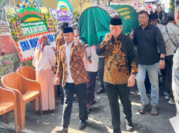 Foto dokumentasi suasana khidmat pemberangkatan jenazah Drs. Sutarto, M.M. di depan rumah duka. Tampak Drs. H. Nurbini, M.S.I. (kiri depan) mengenakan kemeja batik cokelat dan Dr. Bunyamin (kanan depan) mengenakan kemeja batik hitam sedang memikul keranda jenazah hijau bersama jemaah lainnya. Di latar belakang kiri tampak jejeran karangan bunga duka cita.