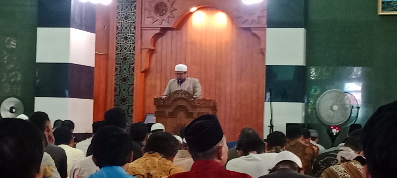 Foto dokumentasi suasana ibadah shalat Jumat di Masjid Roemani Semarang. Tampak khatib Ustaz HM Budi Hidayat, M.Pd.I. mengenakan jas abu-abu, kacamata, dan peci putih sedang menyampaikan khotbah dari atas mimbar kayu berukir di bawah lengkungan kayu bernuansa hangat. Di latar depan, tampak barisan jemaah pria berkopiah hitam sedang duduk tertib menyimak khotbah dari arah belakang. Di sisi kiri terdapat pilar bergaris hitam-putih dan di sisi kanan tampak sebuah kipas angin dinding.