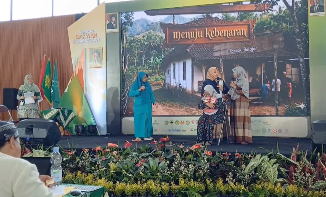 Foto dokumentasi pementasan teater (roleplay) oleh empat orang pengurus perempuan Aisyiyah di atas panggung utama Jambore Dakwah Kemanusiaan. Tampak layar LED raksasa di latar belakang menampilkan visual rumah pedesaan dengan tulisan "menuju kebenaran" dan "Pondok Belajar". Di atas panggung, seorang peserta berbaju biru berdiri memegang mikrofon, sementara di tengah tampak seorang ibu berhijab abu-abu sedang duduk di kursi memeluk boneka anak, didampingi dua pengurus lain yang memberikan pendampingan dengan raut wajah penuh empati