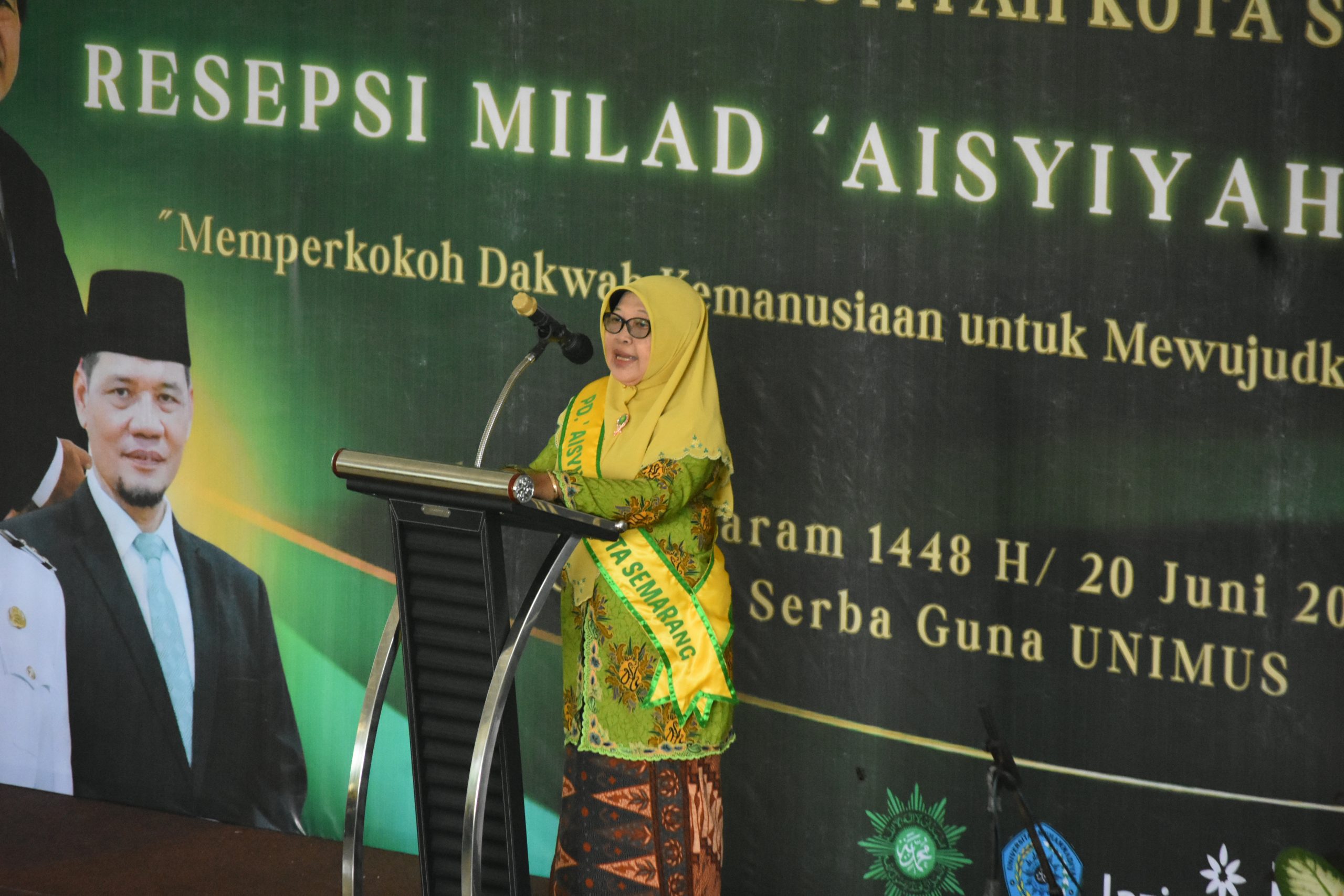 Foto dokumentasi Ketua PDA Kota Semarang Aminah Kurniasih, S.Pd., M.Pd. yang mengenakan kacamata, jilbab kuning cerah, dan busana batik bermotif hijau-emas dengan selempang kuning bertuliskan "PD. 'AISYIYAH KOTA SEMARANG" sedang berbicara secara tenang di balik podium stainless steel. Di latar belakang, tampak sebagian spanduk hijau tua bertuliskan "RESEPSI MILAD 'AISYIYAH" serta sebagian foto tokoh pejabat daerah berjas hitam di sisi kiri.