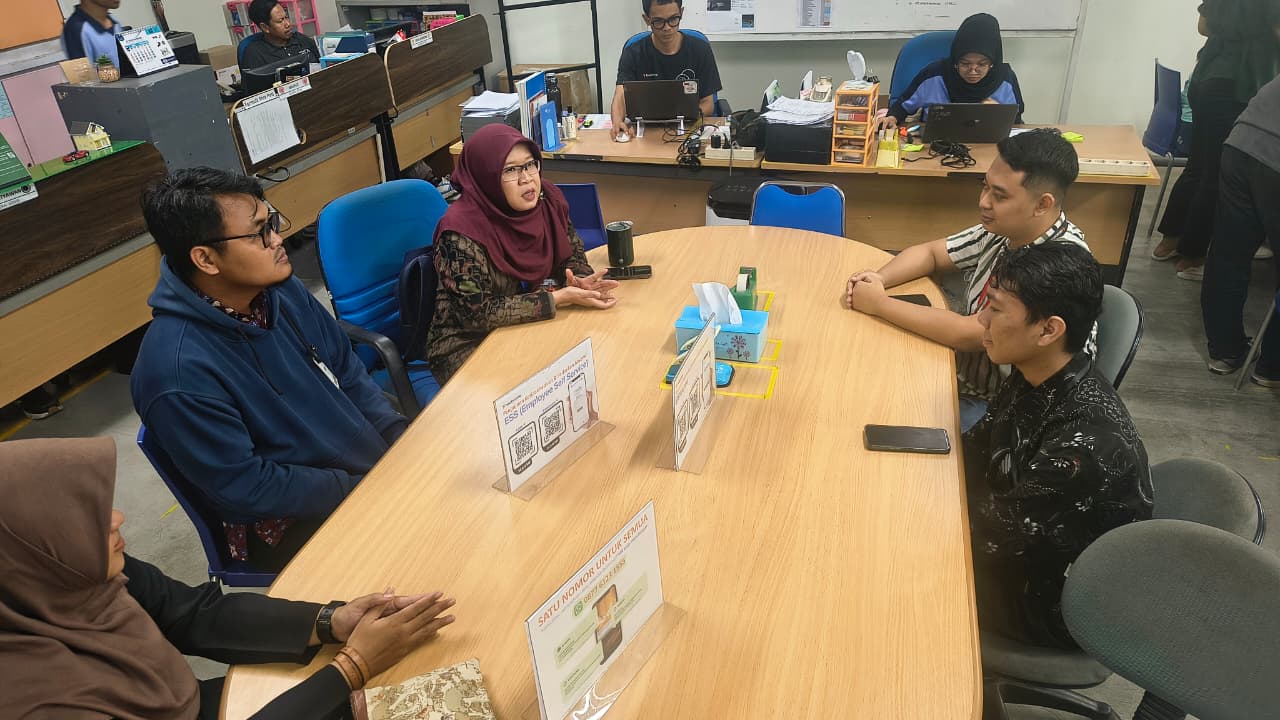 Foto dokumentasi pertemuan evaluasi program magang di sebuah ruang kantor yang kondusif. Tampak sekelompok dosen pendamping dan pihak manajemen PT Apparel One Indonesia duduk mengelilingi meja kayu berbentuk oval. Seorang perwakilan perempuan berhijab marun sedang berbicara dengan ekspresif, sementara peserta rapat lainnya menyimak secara saksama.