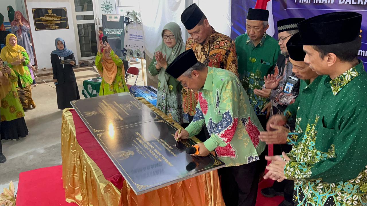 Foto dokumentasi Menteri Pendidikan Dasar dan Menengah RI, Prof. Dr. H. Abdul Mu'ti, M.Ed., yang mengenakan kemeja batik hijau bermotif merah dan peci hitam sedang membungkuk untuk menandatangani prasasti marmer hitam di atas meja berbalut kain merah-emas. Beliau dikelilingi oleh jajaran pimpinan Muhammadiyah pria yang bertepuk tangan khusyuk, sementara di latar belakang kiri tampak beberapa ibu-ibu menyaksikan jalannya prosesi peresmian dengan antusias.