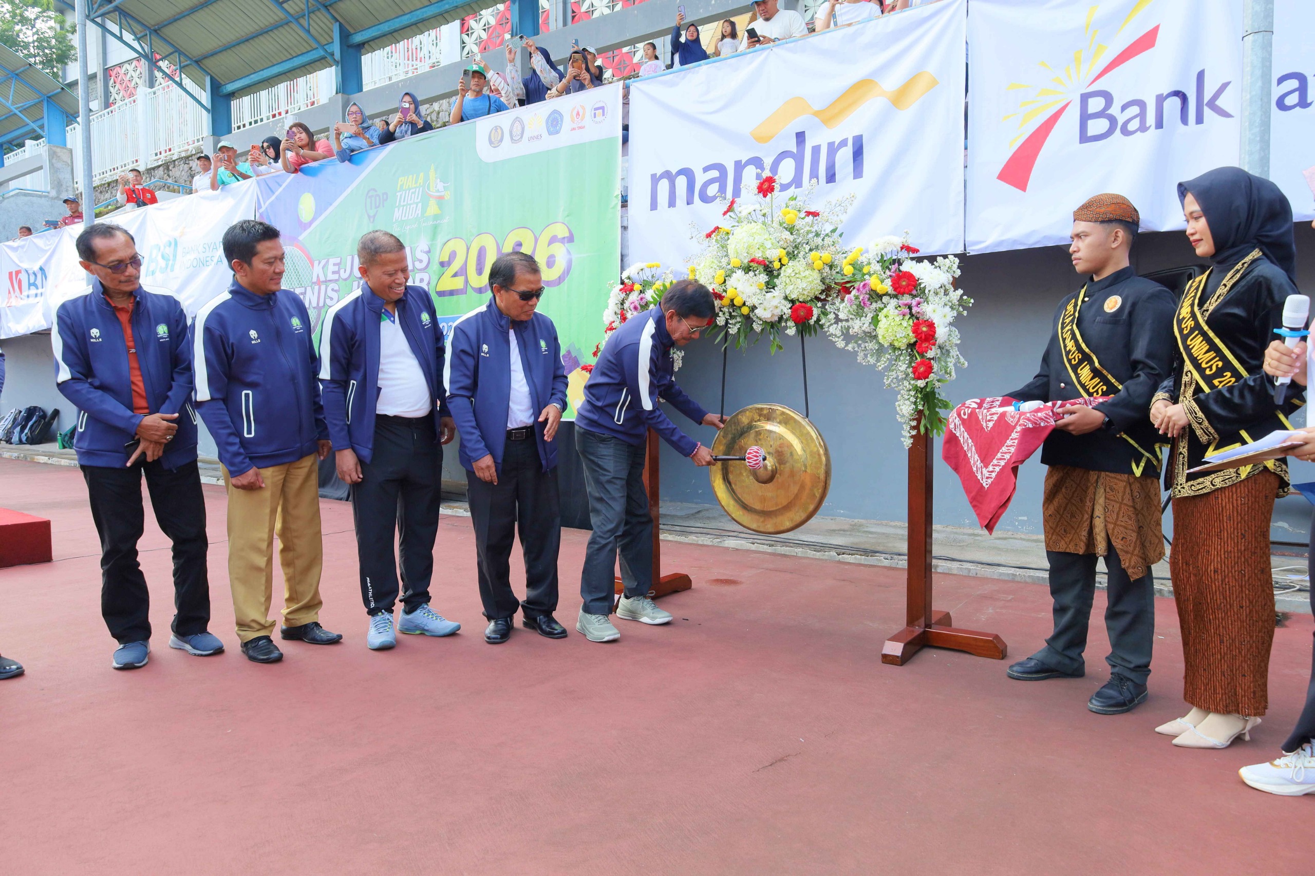 Foto dokumentasi peresmian Kejurnas Tenis Piala Tugu Muda 2026, menampilkan seorang pria berjaket olahraga biru tua sedang memukul gong emas besar yang dihiasi karangan bunga merah-putih. Di sebelah kiri berdiri empat pria berjaket serupa menyaksikan prosesi, sementara di sebelah kanan berdiri dua mahasiswa mengenakan busana adat Jawa sebagai duta kampus UNIMUS.