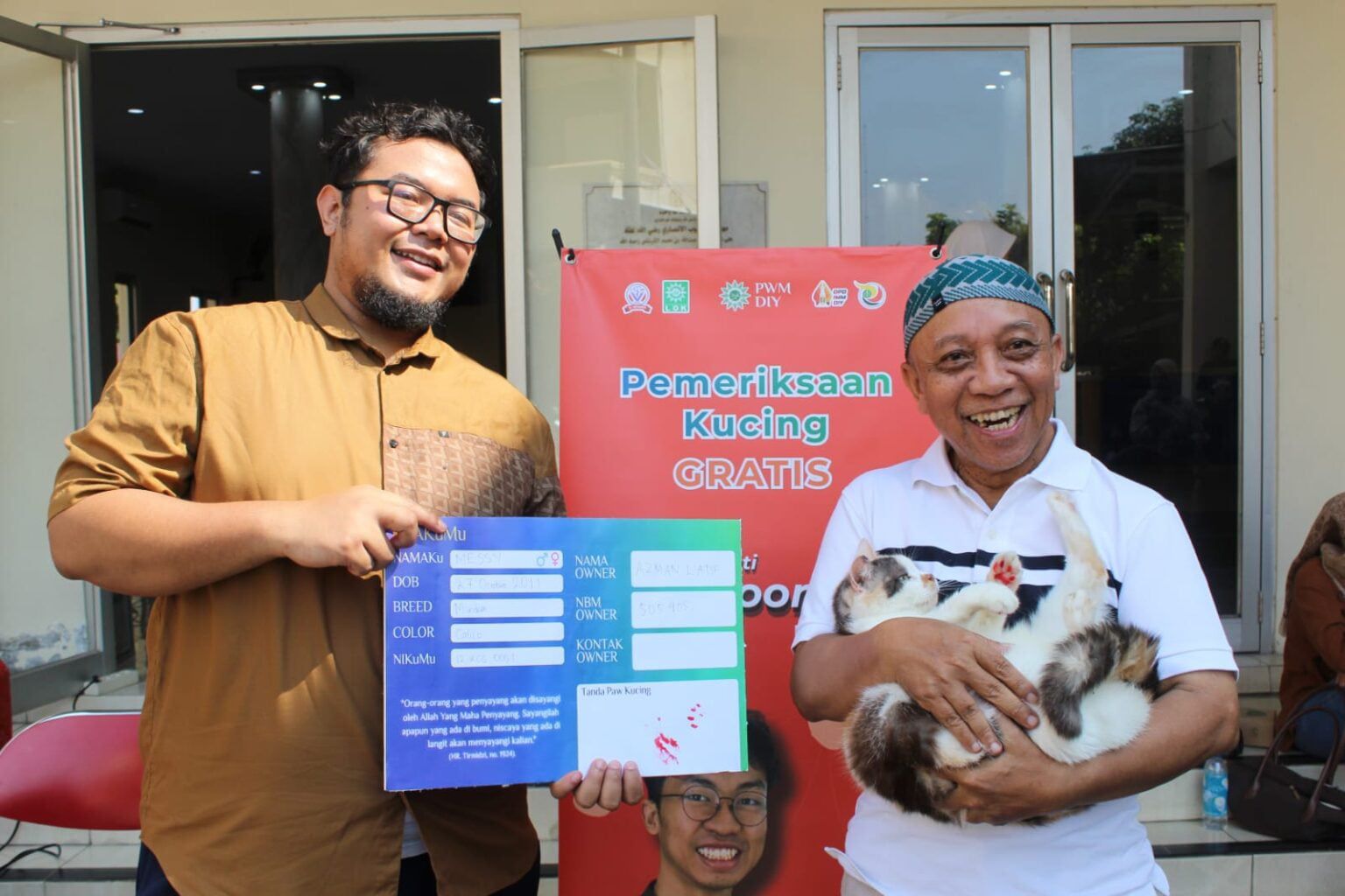 Dua pria tersenyum memegang replika Kartu Tanda Anggota Muhammadiyah (KTAM) KucingMu untuk kucing bernama Messi dan seekor anabul di depan spanduk Pemeriksaan Kucing Gratis PWM DIY.