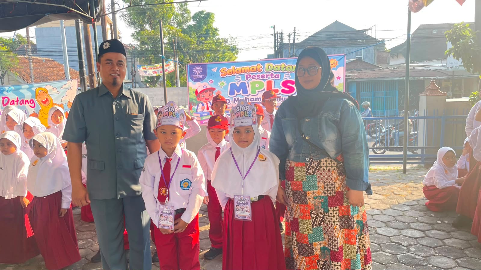 Kepala SD Muhammadiyah 06 Semarang, H. Suharno, dan seorang pendamping menyambut dua siswa baru berseragam merah putih dengan atribut mahkota Siap MPLS dalam pembukaan kegiatan MPLS Ramah Anak di halaman sekolah.
